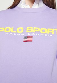 Lila sweatshirt i bomull, med stor gul text "POLO SPORT", "RALPH LAUREN"-logotyp i vitt och en liten design av den amerikanska flaggan.