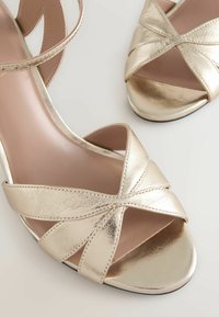Metallic guld højhælede sandaler med krydsede stropper, glat tekstur, åbent tådesign og en slank silhuet.
