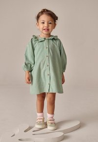 Hellgrünes, knöpfbares Kleid mit Rüschensaum und langen Ärmeln, kombiniert mit beigen und pinken Sneakers sowie rosa Knöchelsocken.