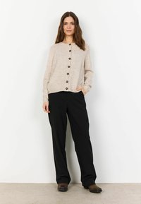Beige pletený cardigan s kulatým výstřihem, zapínáním na knoflíky vpředu a dlouhými rukávy, doplněný o černé kalhoty s širokými nohavicemi a hnědé boty.