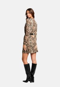 Robe imprimé serpent en beige et marron avec une taille ceinturée. Le modèle porte des bottes noires brillantes et hautes. Texture lisse, silhouette ajustée, longueur au genou.