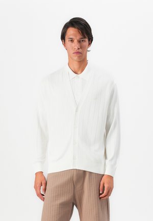 Man wearing white ribbed buttoned cardigan over white collared shirt and beige ribbed trousers, standing against plain white background.