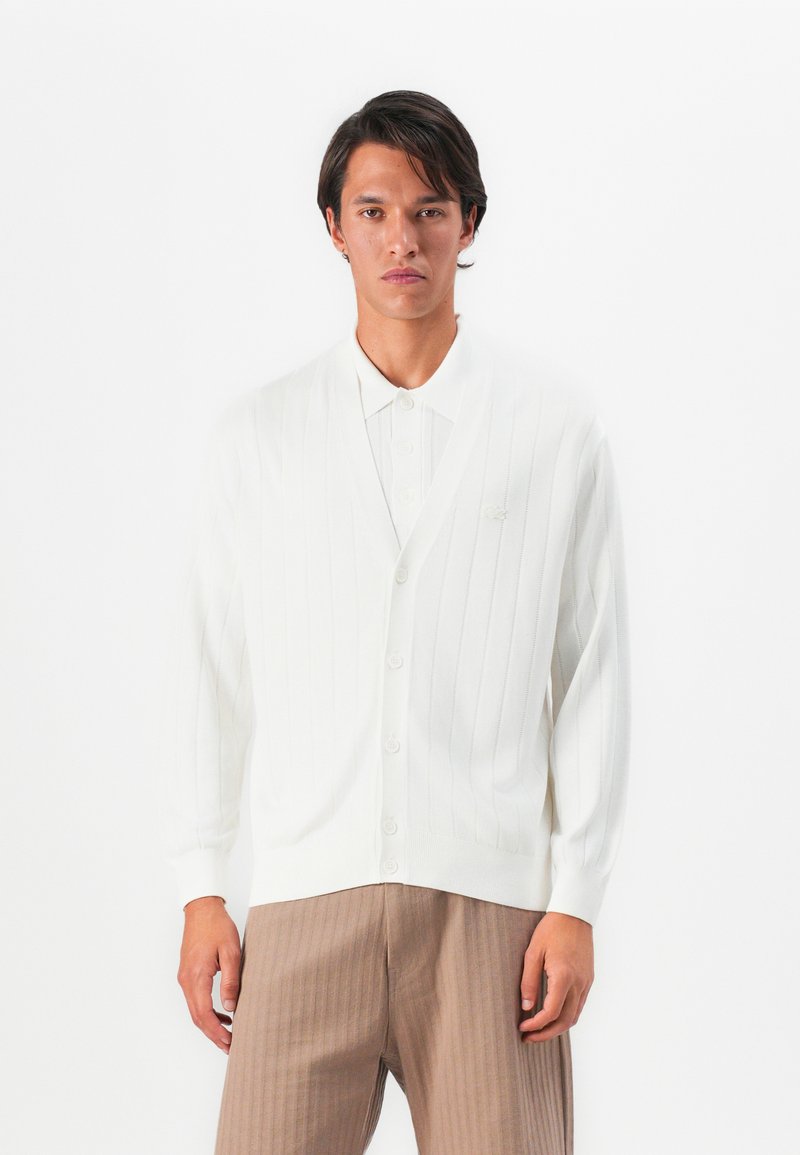 Man wearing white ribbed buttoned cardigan over white collared shirt and beige ribbed trousers, standing against plain white background.