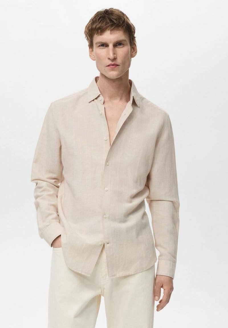 Camicia button-up beige chiaro con una texture in lino, maniche lunghe e un sottile motivo a righe. Indossata con pantaloni bianco sporco.