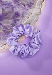 Aksamitna fioletowa gumka do włosów o gładkiej struktury, z zebranym wzorem i małą metką marki, umieszczona na miękkim fioletowym tle.