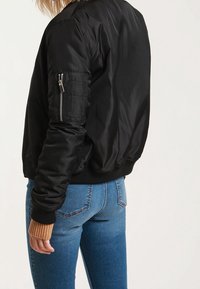 Černá bomber bunda vyrobená z hladké tkaniny, s kapesním zipem na levém rukávu a žebrovanými manžetami. Nosit s modrými džíny.