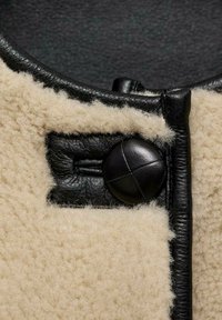 Giacca di pelle nera con dettagli in shearling color crema, dotata di una grande chiusura con bottone nero e texture liscia lungo i bordi.