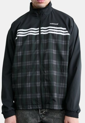 Mand iført sort Adidas lynlåstrøje med hvide striber og gråt ternet mønster foran.