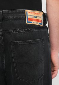 Μαύρο τζιν με υφή. Διαθέτει ορατή πίσω τσέπη και δερμάτινο ετικέτα με μπλε και κόκκινες λεπτομέρειες.