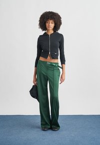 Μαύρο κοντό φούτερ με φερμουάρ και μακριά μανίκια, με μικρό λογότυπο, συνδυασμένο με πράσινο ψηλόμεσο παντελόνι και μαύρα παπούτσια.