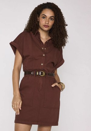 Vrouw met krullend haar die een bruine jurk met knopen en opgerolde mouwen draagt, een donkere riem en gouden armbanden, poserend tegen een effen achtergrond.