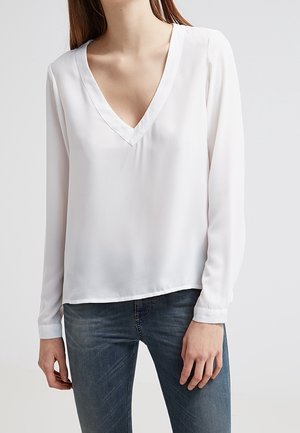 Femme portant un chemisier blanc à manches longues avec un col en V, associé à un jean en denim bleu, debout devant un fond clair uni.