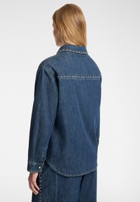 Camicia di jeans a maniche lunghe, di colore blu scuro, con bottoni a scatto ai polsi e borchie metalliche lungo la cucitura della parte superiore della schiena. Texture morbida.