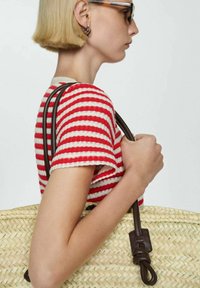 Borsa di paglia intrecciata con manici in pelle marrone. La borsa ha una superficie texturizzata ed è mostrata su uno sfondo neutro. Parte di un top a righe rosse e bianche è visibile.