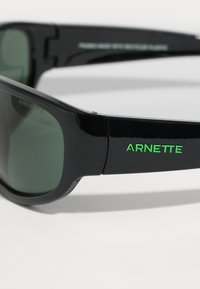 Černé sluneční brýle s zelenými akcenty, s elegantním designem, tmavými skly a značkou "ARNETTE" na boku.