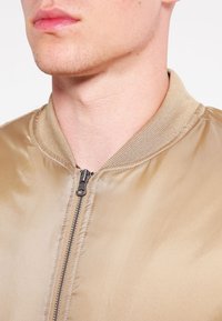 Chaqueta bomber marrón claro con un acabado suave y satinado. Presenta un cuello acanalado y una cremallera central. El diseño es sencillo y minimalista.