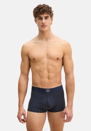 Uomo senza maglietta con capelli ricci che indossa boxer blu navy con pois bianchi, in piedi contro uno sfondo grigio chiaro uniforme.