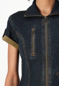 Jeansoverall med hög krage, utrustad med en främre dragkedja, två bröstfickor med dragkedjor och uppvikta ärmar i en kontrasterande färg.