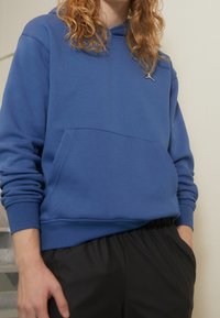 Osoba v modré mikině s kapucí a přední kapsou, v černých kalhotách, stojící uvnitř s rukama částečně v kapsách.