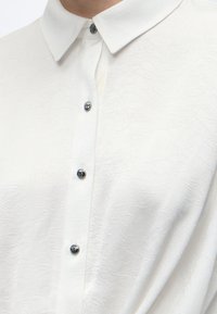 Chemise blanche texturée boutonnée avec col pointu et quatre boutons argentés visibles sur le devant.