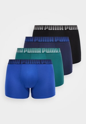 Oben ohne stehender Mann trägt marineblaue Boxershorts mit blauer Bundbeschriftung, steht neben einem abgerundeten hellgrauen Stuhl vor einer einfarbigen hellen Wand.