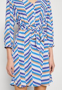 Bunt bedrucktes Wickelkleid mit Zickzackmuster in Blau, Lila und Braun, langen Ärmeln, Knöpfen und einem gebundenen Taillendetail.