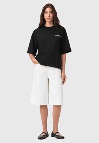 T-shirt nera a maniche corte con vestibilità rilassata e logo bianco, abbinata a pantaloni corti bianchi fino al ginocchio e scarpe nere con dettagli bianchi.