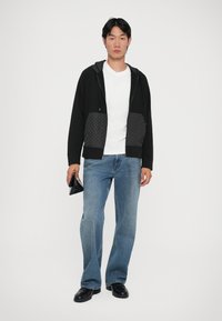 Crna zip-up hoodie s teksturiranim uzorkom na džepu, uparen s svijetloplavim širokim trapericama i crnim cipelama, držeći crnu torbicu.