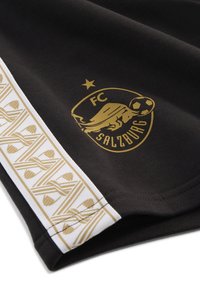 Camiseta negra del FC Salzburg con un logotipo dorado del club, una estrella y una franja blanca con un patrón texturizado dorado a lo largo del lado.