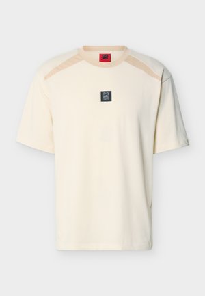 T-shirt crème à manches courtes avec accents beige sur les épaules et un petit patch logo carré noir portant l'inscription "DR HUGO" centré sur la poitrine.