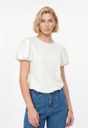Mladá žena s krátkymi blond vlasmi, oblečená v bielom tričku s nafúknutými rukávmi a modrých džínsach, stojaca pred jednoduchým bielym pozadím.