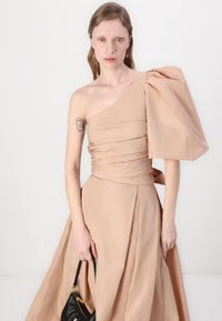 Strapless, beige dress with a dramatic puff sleeve, ruched bodice, and flowing skirt. Model holds a black handbag. Smooth, textured fabric.
