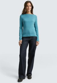 Ein langärmliges, türkisfarbenes Oberteil mit quadratischem Ausschnitt, kombiniert mit dunkelblauen, ausgestellten Jeans. Der Stoff wirkt glatt und eng anliegend.