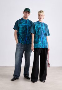 Grafische T-Shirts in Blau und Teal mit abstrakten Mustern. Das männliche Model trägt Jeans, während das weibliche Model sie mit weiten schwarzen Hosen und einer Handtasche kombiniert.