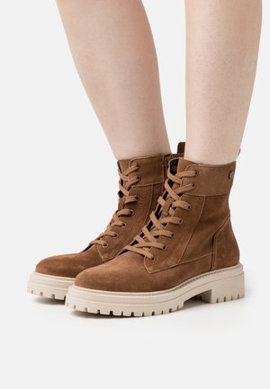 Bruine suède veterlaarsjes met grove beige rubberen zolen, gedragen op blote benen tegen een witte achtergrond.