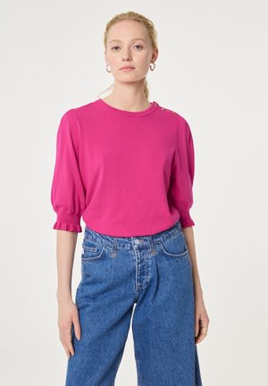 Frau trägt ein leuchtend pinkes Strickoberteil mit Ballonärmeln und Knopfdetails an der Schulter, kombiniert mit hoch taillierten blauen Jeans.