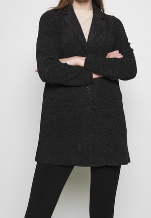 Blazer nero textured con chiusura a un solo bottone e rever a punta, indossato sopra una maglia chiara e pantaloni neri, con maniche lunghe.