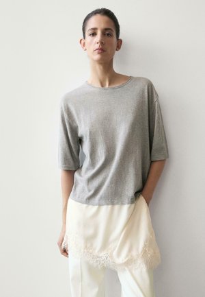 Donna con capelli scuri, indossa una maglia grigia ampia e una gonna bianca con bordi in pizzo sopra pantaloni bianchi, in piedi davanti a una parete chiara e semplice.