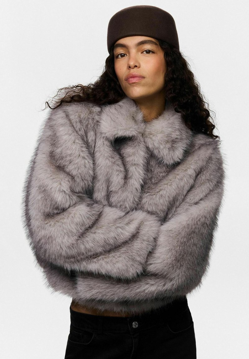 Femme aux cheveux bouclés portant un chapeau marron et un manteau en fausse fourrure gris, les bras croisés, regardant la caméra avec confiance sur un fond uni.