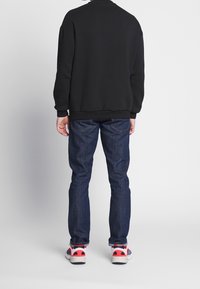 Persona con una felpa nera, jeans blu scuro e scarpe bianche con accenti rossi e blu marino, in piedi di spalle su uno sfondo neutro.