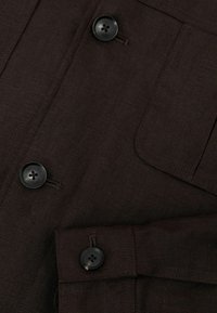 Tela texturizada de color marrón oscuro con tres botones negros de cuatro agujeros, un bolsillo en el pecho y un puño abotonado visible en vista de primer plano.