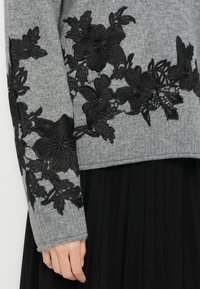 Pull en tricot gris avec des appliqués en dentelle florale noire sur les manches et la taille, associé à une jupe plissée noire.