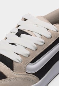 Κοντινό πλάνο σε μπεζ, μαύρο και λευκό αθλητικό παπούτσι με πλατιά λευκά κορδόνια και εμφανείς ραφές σε σουέτ και δίχτυ υλικά.