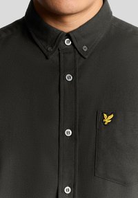Camisa de botón de color verde oscuro con un tejido texturizado, que presenta un logo amarillo bordado en el bolsillo del pecho y botones plateados.
