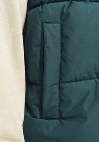 Groene puffer gilet met een gladde textuur, voorzien van een patchzak aan de zijkant. Het materiaal is lichtgewicht en strak, met een gequilt ontwerp.