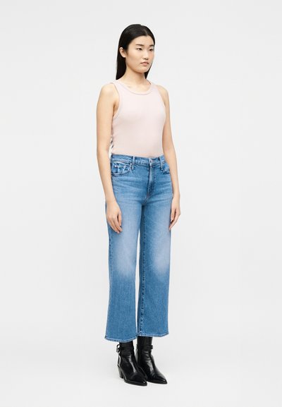 Γυναίκα που φοράει ένα ανοιχτό ροζ αμάνικο τοπ, ψηλόμεσα κοντά μπλε τζιν και μαύρα μποτάκια στον αστράγαλο, στέκεται μπροστά σε ένα απλό φόντο.