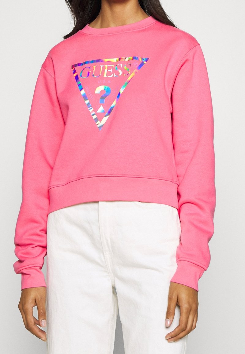 Femme portant un sweat-shirt rose vif à manches longues avec un logo Guess holographique et un pantalon blanc taille haute, debout devant un fond uni.