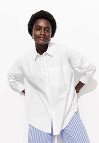 Chemise blanche à boutons avec une seule poche poitrine, manches longues, texture douce et coupe décontractée, associée à un pantalon rayé bleu.