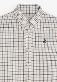 Camisa de cuadros en blanco, con patrones en cuadrícula en azul oscuro y óxido, y un pequeño logo de calavera bordado en el pecho.