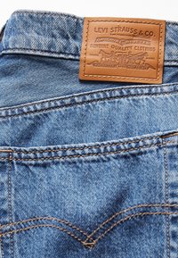 Jean en denim bleu clair avec une étiquette en cuir marron Levi Strauss & Co. présentant le logo classique des deux chevaux et des détails de couture.
